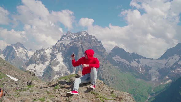 Young Man Sitting on Rock and Taking Photos of Mountain Landscape on Smartphone alt