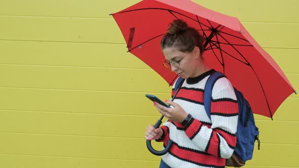 Girl Holds Red Umbrella Looks to Phone Types alt