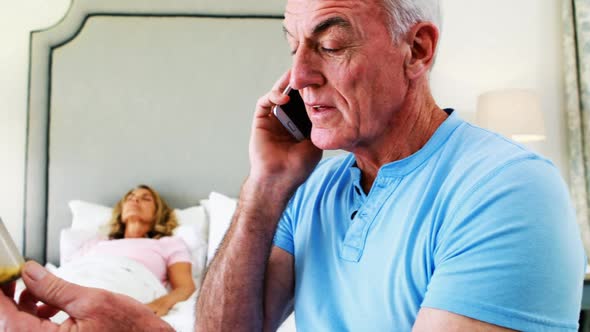 Senior man talking on mobile phone while senior woman resting in bedroom alt