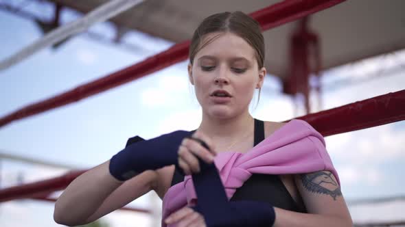 Confident Focused Female Boxer Wrapping Wrist with Boxing Bandage Sitting on Ring Outdoors alt