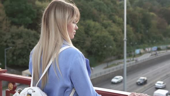 Business Girl Stands on a Pedestrian Bridge and Looks at the Stream of Cars alt