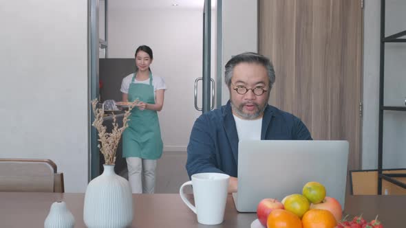 Middle age man working on laptop and wife prepares meal and serve to husband