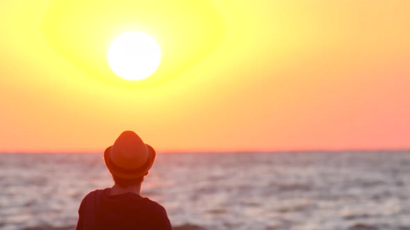 Young Guy in a Hat Watching the Sunset By the Ocean. The Male Traveler Enjoys the View of the East