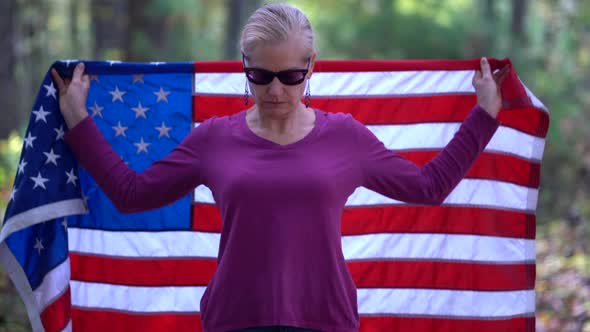 Blonde woman raising an American flag behind her with serious expression. alt