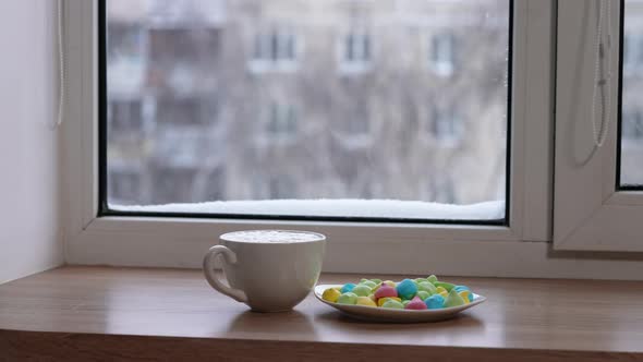 A Large Mug of Coffee with Milk or Mochacino and Colorful Sweets in a Plate on the Windowsill in