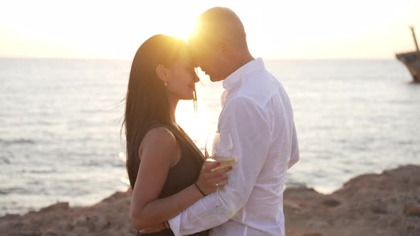 Side View Happy Loving Couple Dancing in Sunbeam on Mediterranean Sea Beach with Wine Glasses alt