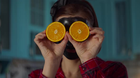 30s woman with black cosmetic mask on her face holding orange fruit halves near eyes alt