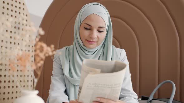 Muslim Woman Wearing Traditional Hijab Reading Newspaper at Table in Cafe alt