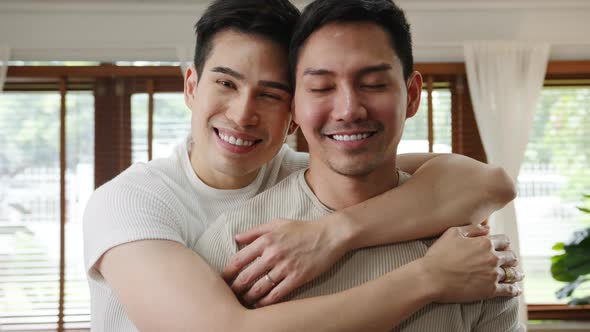 sian LGBTQ men relax toothy smile looking to camera while hug in living room .