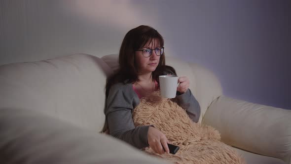 Brunette Pretty Woman Sits on the Couch with a Cup of Tea in the Evening and Watches Tv