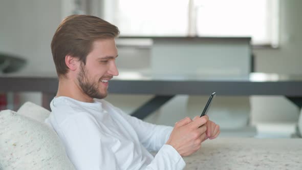 Cheerful Caucasian Man Sits at Home on the Couch and Uses a Smartphone Surfing Internet Pastimes alt