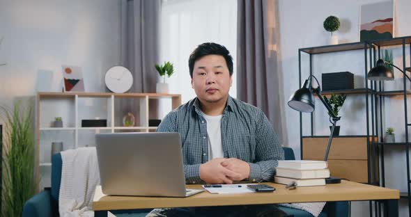 Asian Man Sitting in Front of Camera at His Workplace in Home Office alt