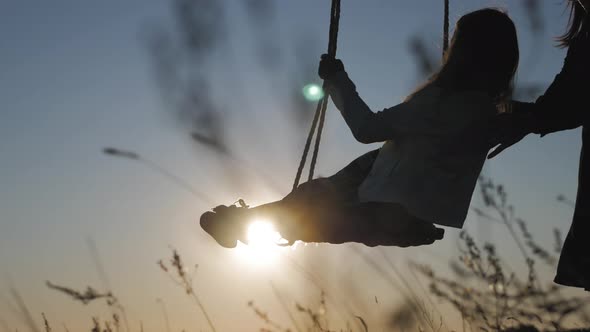 Silhouette of Happy Young Mother and Little Daughter on a Swing at Sun Light alt