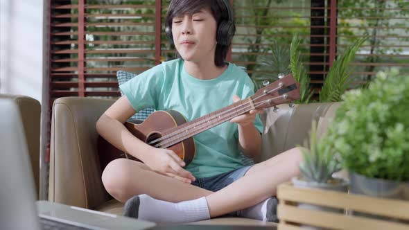 Little asian child boy online learning playing ukulele on armchair at living room