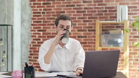 Pretty Woman with Short Haircut and White Shirt Drinks Coffee From a Paper Cup Typing on Laptop and alt