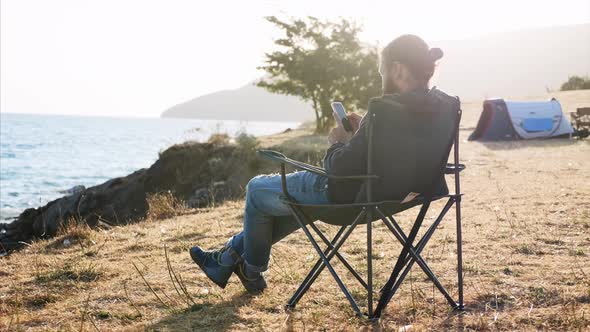 Young Man Sitting on Folder Chair and Using Smart Phone at Camping at Lake Shore