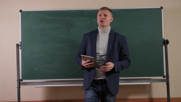 A Young Male Teacher Stands in the Lecture Room with a Tablet in His Hands alt