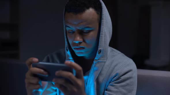 Unhappy African-American Teenage Procrastinator Losing Video Game on Smartphone alt