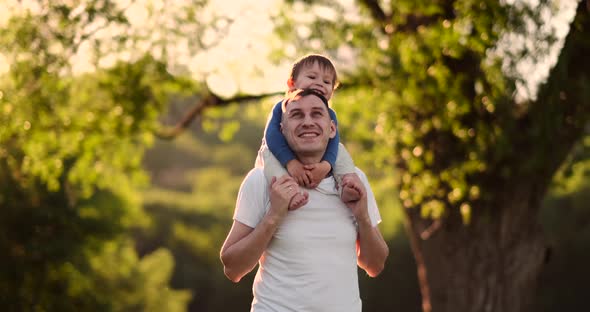 A Child Sitting on the Neck of His Father While Walking in Summer Field at Sunset alt