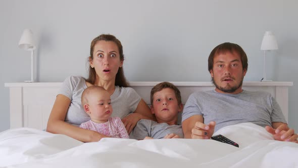 Happy Young European Family with Two Kids Gather on Couch to Watch TV in the Badroom Smiling alt