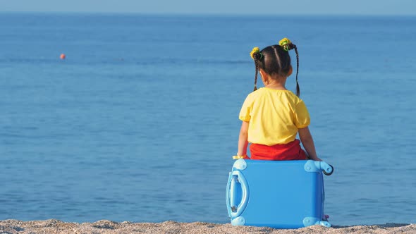 Little Cute Girl in Colorful Clothes Sitting on a Blue Suitcase on the Sea Beach. alt