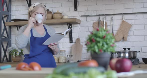 Portrait of Mature Blond Woman Drinking Coffee and Reading Book of Recipes in the Morning. Serious alt