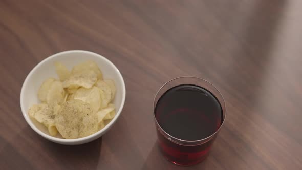 Slow Motion Handheld Shot of Potato Chhips with Red Drink in Tumbler Glass on Walnut Table with Copy alt