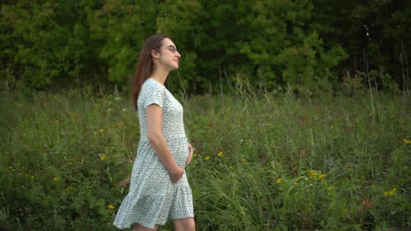 Happy Young Pregnant Woman Walks in Nature and Strokes Her Belly alt