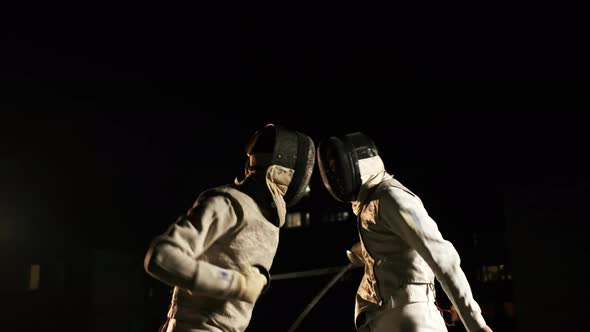Two Fencers In Fencing Suits And Helmets Fencing In The Dark Outside. alt