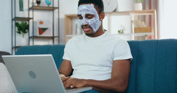 Black-Skinned Guy with Face Mask Sitting in front of Laptop and Chatting with Friends alt