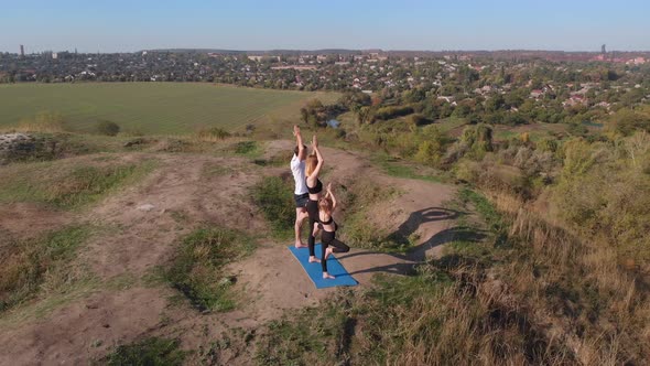 Orbiting Aerial Shot of Family of Three Mother Father and Daughter Do Yoga Exercises on Top of Hill alt