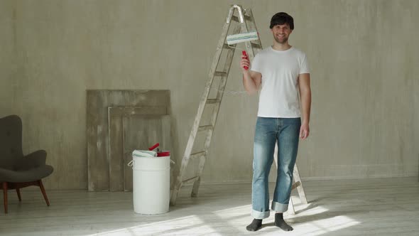 Young Man Holding a Roller Against the Background of a Bucket of Paint and a Ladder alt