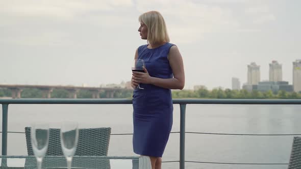 Thoughtful Charming Lady in Blue Dress Standing in Outdoor Cafe on River Bank with Wineglass alt