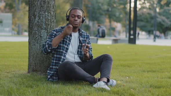 Afro American Guy Sits in Park on Grass in Summer Day Listening Music on Headphones Sings Song alt