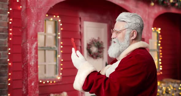 Santa Claus Communicates Via Video Link with Someone Using Smartphone on the Background a Red House