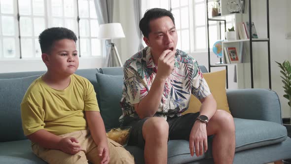 Asian Father And His Son Are Watching Tv While Eating Potato Chips At Home alt