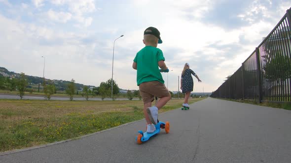Little Boy Rides Three-wheeled Scooter and His Older Sister on Skateboard Outdoor in the Park alt