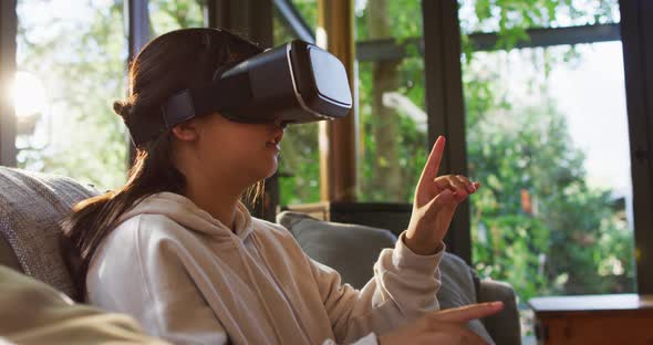 Asian girl smiling and gesturing while wearing vr headset sitting on the couch at home alt