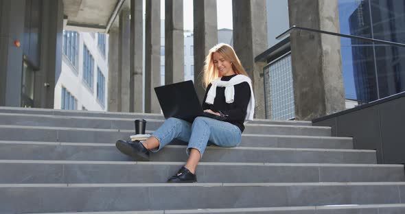 Girl in Casual Clothes Sitting on Steps of Office Building and Uses Her computer alt