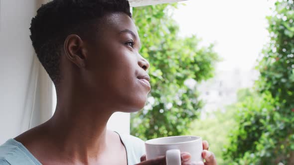 African american woman holding coffee cup looking out of window at home alt