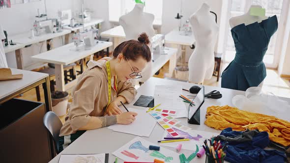 Young Woman Fashion Designer Watching Online Webinar on Digital Tablet and Drawing Sketches at alt