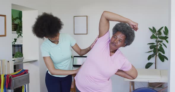 Happy african american female physiotherapist helping senior female patient exercise at home alt
