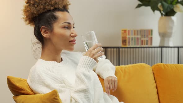Young Black Woman Drinking a Glass of Water Sitting on the Sofa in the Living Room alt