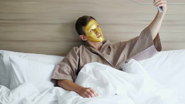 Guy with Golden Face Mask Makes Selfie in Soft Bed in Hotel alt
