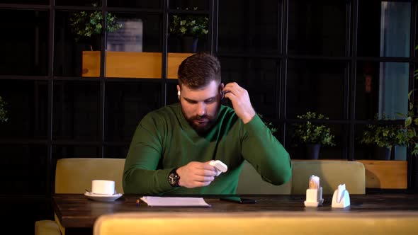 Bearded Hipster in Bluetooth Headphones See Important Work Papers in the Cafe