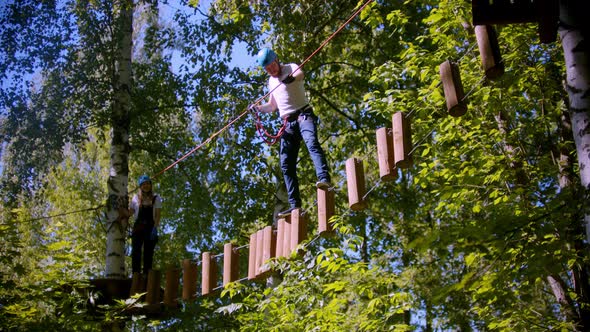 A Man Walks on Logs Suspended in the Air Between Trees in the Forest - a Woman Waits for Him To alt