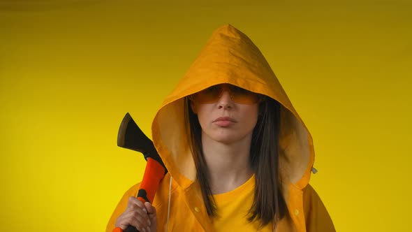 Young Woman in a Yellow Raincoat Holds an Axe in Her Hand in a Yellow Studio alt