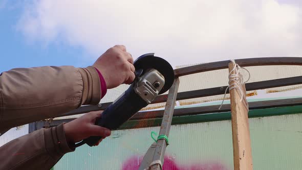 A Man with an Angle Grinder Cuts a Rusty Metal Greenhouse Against a Blue Sky alt