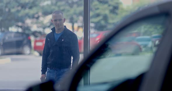Young Man Looks at the Car Through the Showcase of a Car Dealership alt