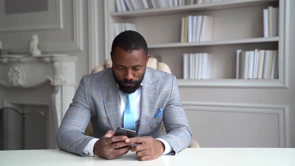 Tired Rich Man Is Checking News in Internet By Mobile Phone Sitting at Table in His Home, Leaning on alt